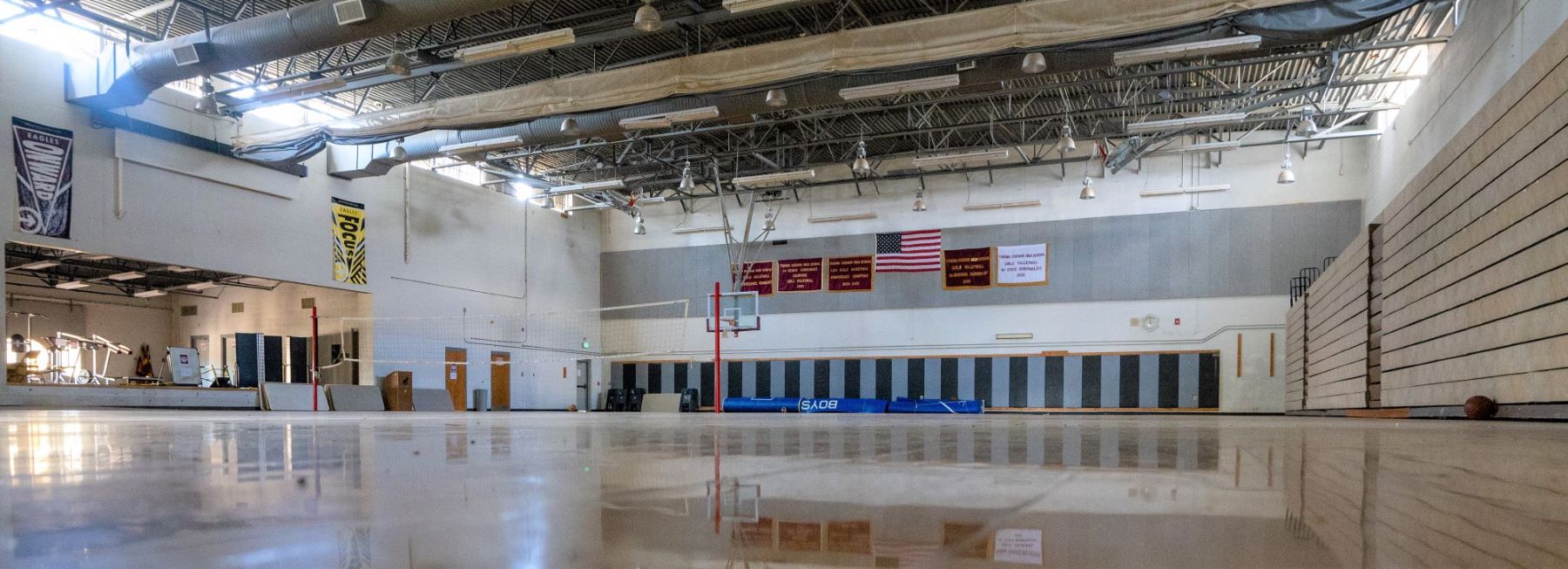 A spacious school gymnasium featuring a basketball hoop and a flag hanging on the wall.