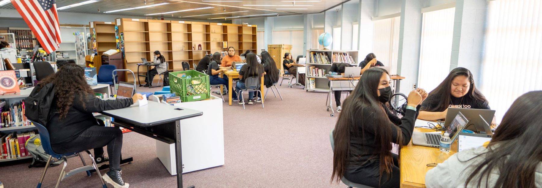 Students collaborate on laptops while seated at tables in a busy library.