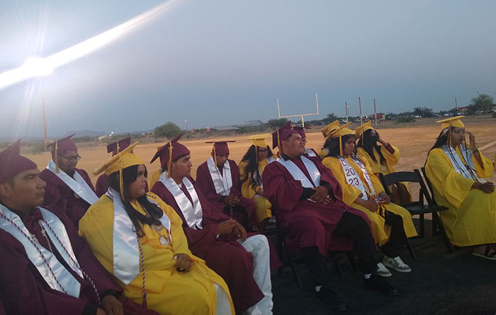 Students seated outdoors wearing red and yellow graduation caps and gowns.