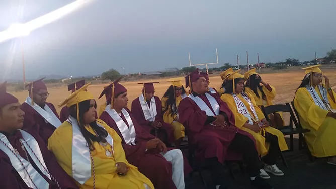 Students seated outdoors wearing red and yellow graduation caps and gowns.